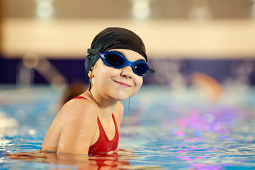 Girl child swimmer in a red bathing suit on a background of the pool inside the room with glasses and a cap