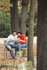 Young couple reading guidebook on park chairs along tree trunks