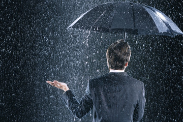 Rear view of a businessman staying dry under umbrella during downpour - obrazy, fototapety, plakaty
