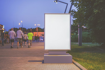 Summer evening. Front view. Blank vertical bilbor stands in city park. In the background, a walking group of people. Mock up. Advertising space.