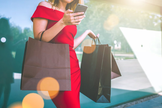 Cropped image.Young smiling woman with blond hair,dressed in red fitting dress,standing outside with shopping bags and using smartphone.Girl uses digital gadget. Summer day.On background glass wall.