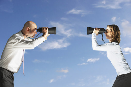Side view of two business colleagues looking at each other through large binoculars against sky