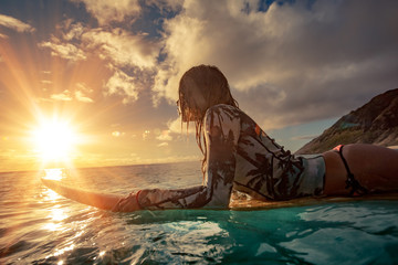 A surfer girl watching sunset on a surboard floating in blue ocean near rocky shore - Powered by Adobe