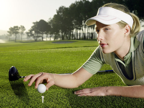 Beautiful young female golfer lying on grass while placing ball on tee - Powered by Adobe