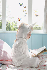 Side view of a young girl reading book in unicorn costume on bed