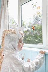 Profile of a smiling young girl in unicorn costume looking at artificial butterflies on window