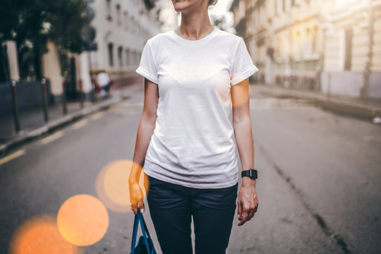 Summer day. Front view. Young woman in a white T-shirt and blue pants standing outside. In the background, the city street. Cropped image. Bokeh effect. Mock up.
