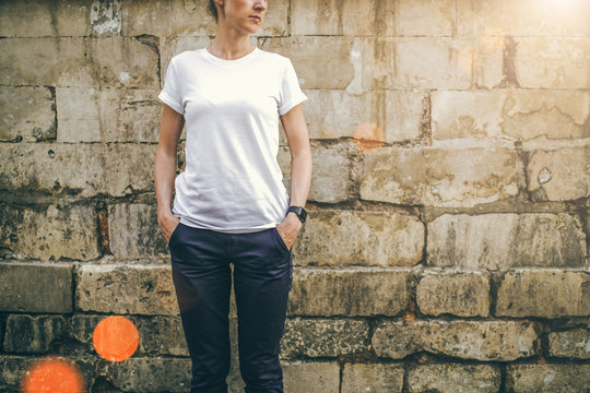 Summer day. Front view. Young woman in a white T-shirt and blue pants standing on a background of a stone wall with his hands in his pockets. Cropped image. Mock up.