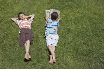 Elevated view of a boy reading besides asleep brother on grass