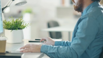 Young Bearded Man Taking Notes while Working on Desktop Computer. He Sips Coffee. He Sits in a Creatively Designed Office.  Shot on RED Cinema Camera in 4K (UHD).