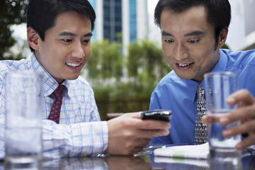Two young businessmen reading text message on cell phone at outdoor cafe