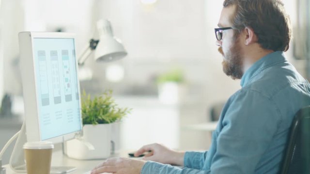 In the Creative Agency Office. Bearded Man in Glasses Works at the Desktop Computer. Camera Moves From His Hands To a Close-up of His Face and Computer Screen. Shot on RED Cinema Camera in 4K (UHD).