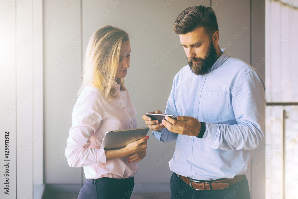 Wall mural Businesswoman and bearded businessman standing in an office near window and discuss business plan.Man showing woman information on smartphone.Girl holding tablet computer.Teamwork. Businesspeople.