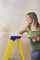 Smiling young woman leaning on step ladder with mug against wall