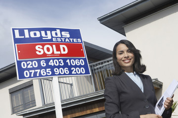 Portrait of a happy and confident female real estate agent standing by sold sign outside house