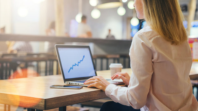 Rear view. Young business woman dressed in light pink shirt sitting in cafe at wooden table and using laptop. On desk is smartphone and cup of coffee. Girl browsing internet, working online.