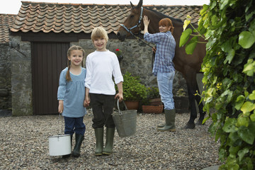 Portrait of sister and brother holding buckets with mother and horse by stable