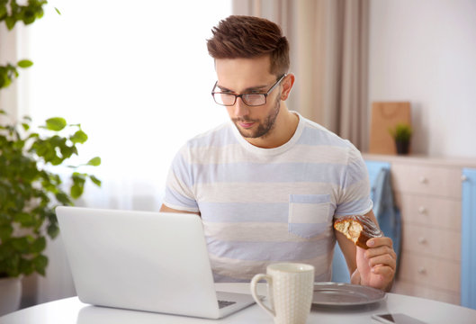 Young man having snack while working with laptop in kitchen