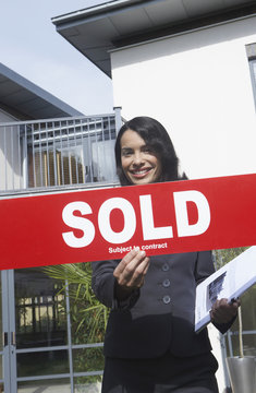 Portrait Of A Smiling Female Real Estate Agent Holding Sold Sign Outside House