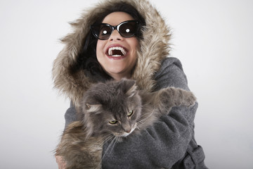 Laughing young woman in hooded fur coat holding cat on gray background