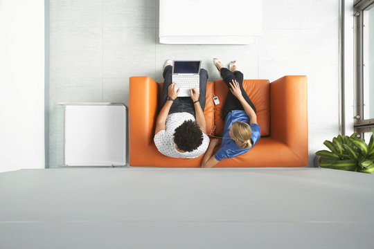 Full Length Top View Of A Man And Woman Using Laptop On Orange Sofa In Reception Room At Office