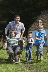 Full length of cheerful father and children racing with wheelbarrow outside cottage