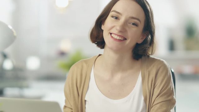 Focused Young Woman Stops Working on Her Laptop and Smiles at the Camera. Her Background is Blurred.  Shot on RED Cinema Camera in 4K (UHD).
