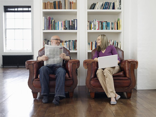 Full length of a middle aged couple sitting in armchairs with newspaper and laptop at home