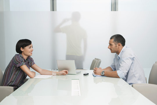 Side view of a man and smiling woman talk over conference table with man on cellphone across translucent office wall