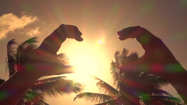 CLOSE UP: Making heart with hands over rising sun with lush palms in background