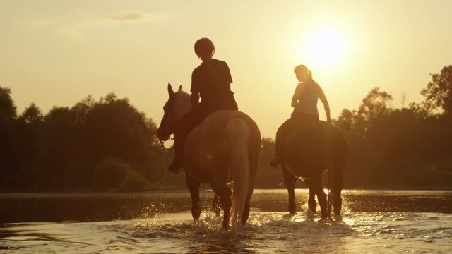 CLOSE UP: Two girls horseback riding horses along the river at magical sunset