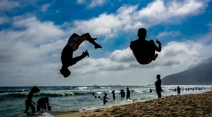 Silhouettes of two men performing somersaults at sand of Ipanema beach with silhouettes of swimming...