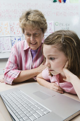 Teacher helping schoolgirl use laptop in classroom