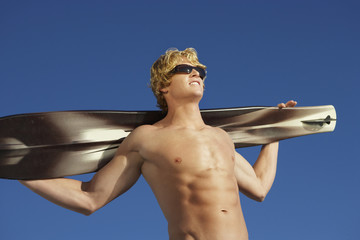 Low angle view of young man holding wakeboard against blue sky