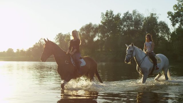 CLOSE UP: Two young girls crossing wide river on horses on stunning sunny day
