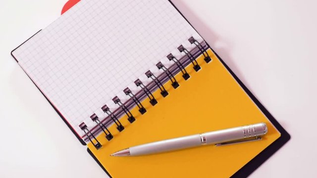 Silver pen lying on top of notebook with tabs, orange tab open, white background. Close up, top view, seamless loop, rotation, 4K Ultra HD.