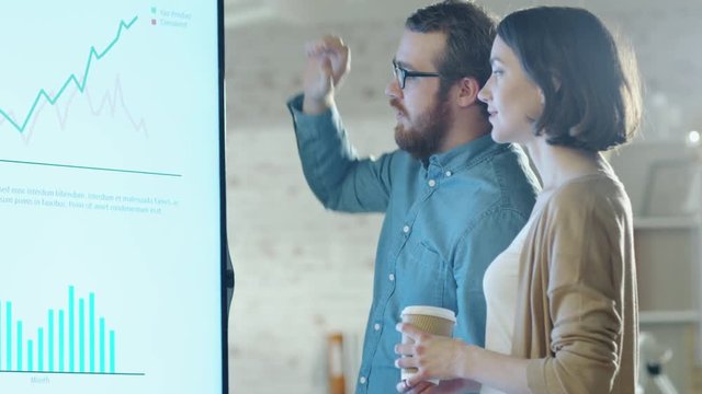 Young Man and Woman Discuss Charts Drawn on Their Electronic Whiteboard. Man Shows Details on the Screen Woman Listens Holding Cup of Coffee in Her Hands. Shot on RED Cinema Camera in 4K (UHD).