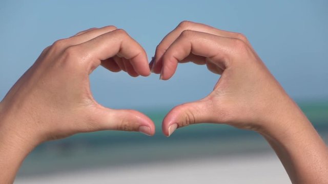 CLOSE UP: Making heart symbol with hands over blue cloudless sky and emerald sea