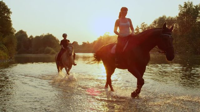 CLOSE UP: Young girls riding horses by the riverbank at golden sunrise