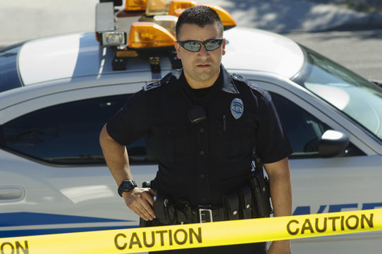 Male Police Officer Standing By Car Behind Caution Tape