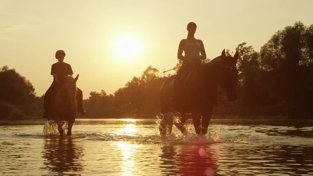 CLOSE UP: Palomino and dark brown horse with riders in river at magical sunset