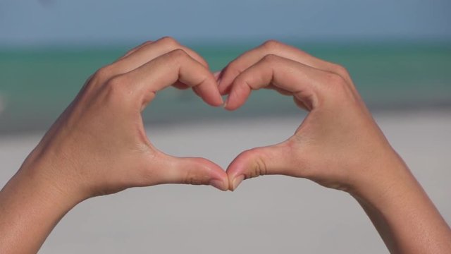 CLOSE UP: Making heart with hands over blue sky, emerald sea and sandy beach 