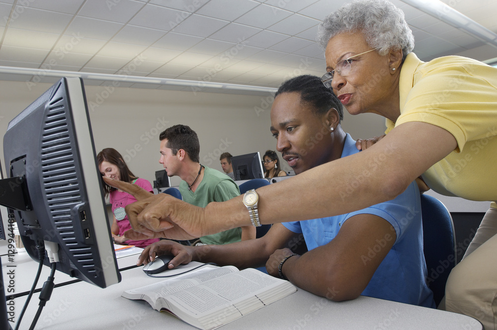 Wall mural senior teacher assisting male student during computer class with classmates in the background