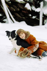 Young girl child sitting on the snow in the winter in a sheepskin coat and hugging dog