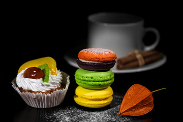 Brightly lit foreground with cake, macaroons in stack and dry orange physalis gradually moving to dark background with wihte cup for coffee and cinnamon rolls on saucer