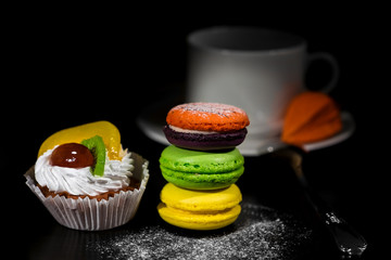 Brightly lit foreground with cake, macaroons in stack gradually moving to dark background with wihte cup for coffee on saucer with spoon and  orange dry physalis on it