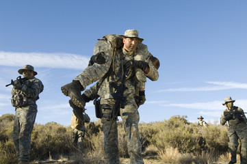 US military man carrying injured female solider on shoulder with teammates in the background
