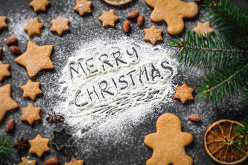 Obraz na płótnie Canvas Merry Christmas greeting written on flour. Christmas holidays card with gingerbread cookies, spices, fir tree. Selective focus