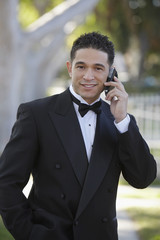 Portrait of a happy young Hispanic man in tuxedo using cell phone outdoors at Quinceanera