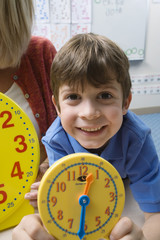 Portrait of a preadolescent boy showing yellow clock with teacher in the background